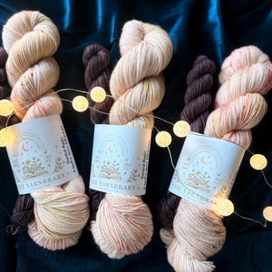 May include: Three skeins of cream-colored yarn with subtle pink and gold speckles, each paired with a smaller dark brown mini-skein. Each set features a white paper label printed with The Yarnbrary logo, text, and Practical Magic branding.