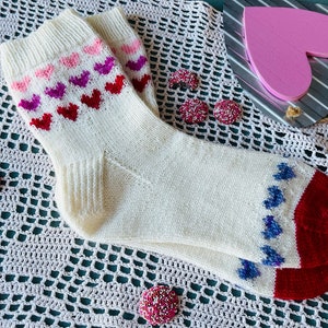 May include: White knitted socks with a red, pink, and purple heart pattern on the top and blue hearts on the heel and toe. The socks are on a white crocheted surface.