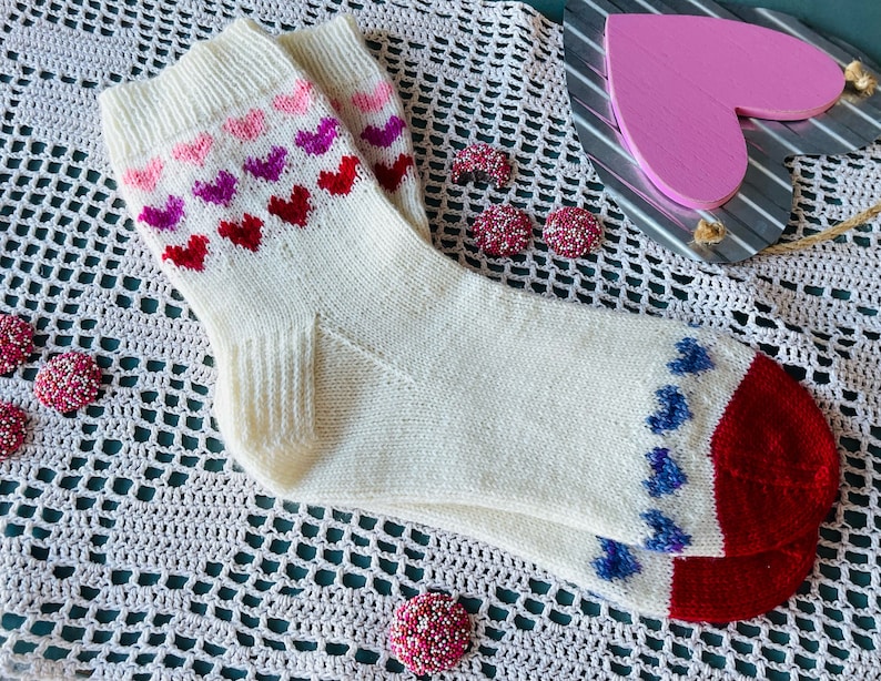 May include: White knitted socks with a red, pink, and purple heart pattern on the top and blue hearts on the heel and toe. The socks are on a white crocheted surface.