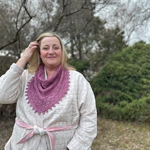 Może przedstawiać: Kobieta w białej kurtce z różowym szalem na szyi. Szal jest wykonany na szydełku i ma koronkową krawędź.