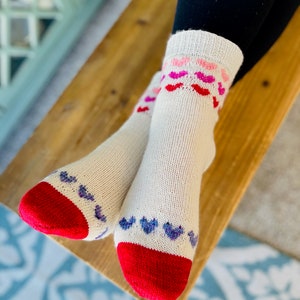 May include: A pair of white socks with red heels and toes. The socks have a pattern of pink, red, and purple hearts on the top of the foot.