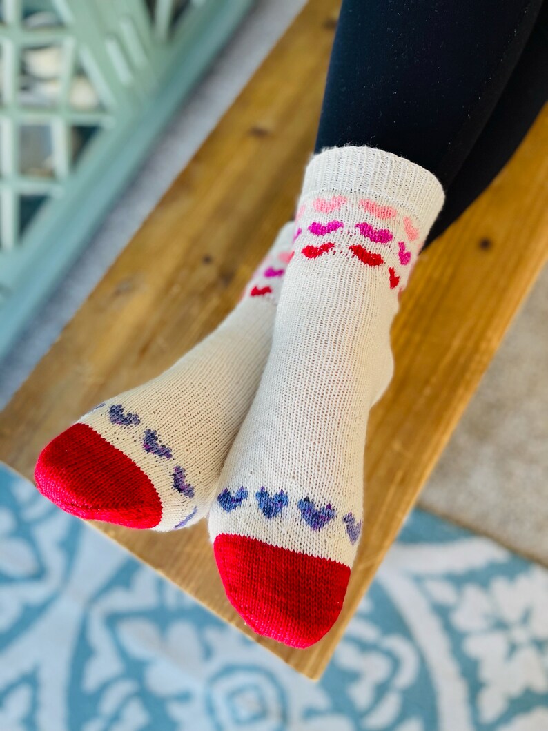May include: A pair of white socks with red heels and toes. The socks have a pattern of pink, red, and purple hearts on the top of the foot.