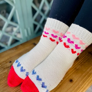 May include: A pair of white knitted socks with a red and blue heart pattern. The socks have a red cuff and a white background.