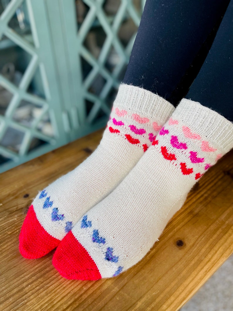 May include: A pair of white knitted socks with a red and blue heart pattern. The socks have a red cuff and a white background.