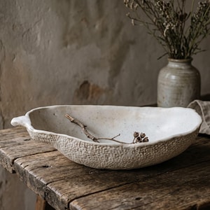 Może przedstawiać: Płytka, złamane bieli ceramiczna miska o nieregularnym, organicznym kształcie i teksturowanej, ziarnistej powierzchni zewnętrznej. Wnętrze jest gładkie z subtelnymi ciemnymi plamkami, a wewnątrz leży mała, cienka, brązowa gałązka.