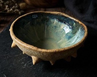 Puede incluir: Un cuenco de cerámica hecho a mano con un diseño texturizado único. El cuenco presenta un exterior moteado, de color beige y un interior brillante azul-verdoso. El borde es ligeramente irregular y la base tiene varias pequeñas protuberancias puntiagudas. El cuenco se muestra sobre una superficie oscura.