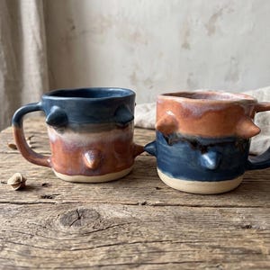May include: Two handmade ceramic mugs with rounded handles and three protruding conical spikes on the exterior. The left mug features a dark blue top fading into a reddish-brown base, while the right mug has a reddish-brown top and dark blue base.