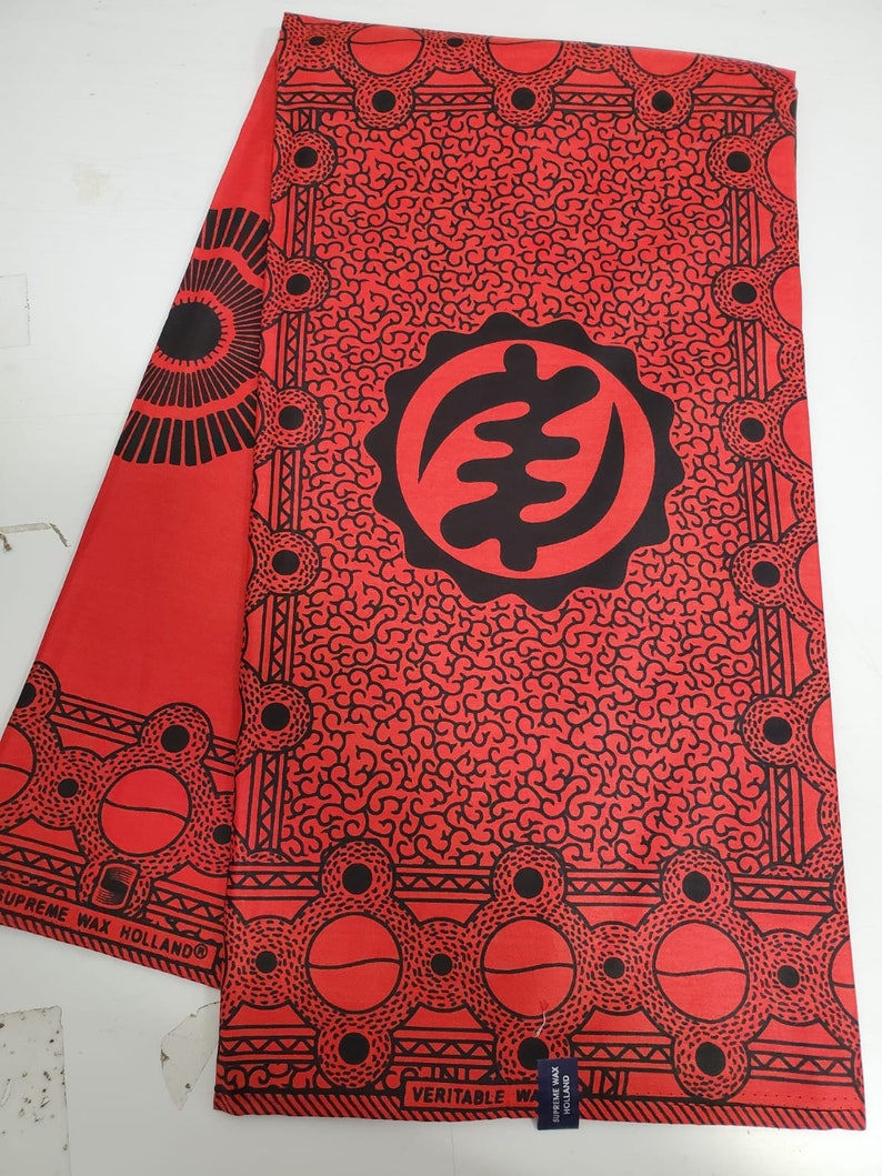 May include: Red fabric with a black and white pattern. The pattern features a large circle with a symbol inside, surrounded by smaller circles. The fabric has the text "SUPREME WAX HOLLAND" and "VERITABLE WAX HOLLAND" printed on it.