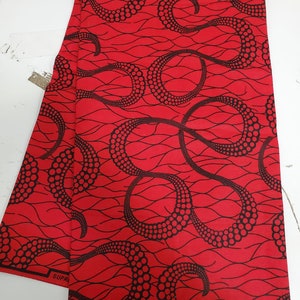 May include: Red fabric with a black pattern of swirling lines and dots. The fabric has the text "SUPREME" and "VERITABLE WAX" printed on it. The fabric also has a label that says "HOLLAND" and "MADE IN NIGERIA".