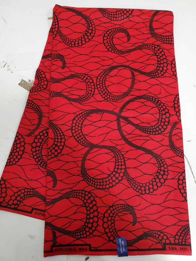 May include: Red fabric with a black pattern of swirling lines and dots. The fabric has the text "SUPREME" and "VERITABLE WAX" printed on it. The fabric also has a label that says "HOLLAND" and "MADE IN NIGERIA".