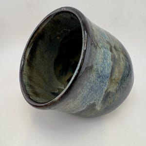 May include: A ceramic mug with a rounded, bulbous shape and a wide, open rim. The exterior and interior feature a reactive glaze with blended tones of deep navy blue, charcoal gray, and muted sage green, creating a smooth, glossy finish.