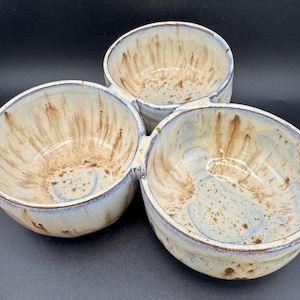 May include: A ceramic serving dish featuring three interconnected round bowls. The pottery has a cream-colored base glaze with brown drip patterns and blue-tinted rims. The interior of each bowl shows a speckled texture with radiating brown streaks.