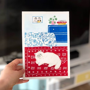 May include: A watercolour illustration on paper depicting a white fluffy cat lying on a red patterned rug. Above the rug, a blue dresser holds a potted plant and books labelled "WARBLES," "LIVING BUILDINGS," and "BRICK." A framed portrait hangs on the wall.