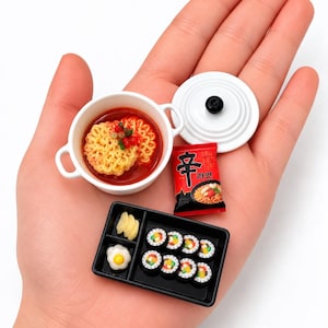 Könnte beinhalten: Ein Miniatur-Kochtopf in Weiß mit Deckel, gefüllt mit roter Brühe und gelben Nudeln, eine kleine rote Packung mit dem Schriftzeichen 辛 und koreanischem Text sowie ein schwarzes rechteckiges Tablett mit acht Sushi-Rollen, Ingwer und einem Spiegelei.