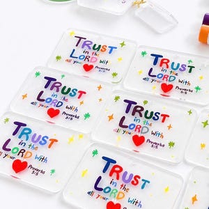 Può includere: Portachiavi rettangolari trasparenti con la scritta "TRUST in the LORD with all your heart Proverbs 3:5" in lettere colorate, un cuore rosso e accenti di stelle e fiori. Un portachiavi ha un anello di metallo verde.