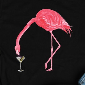 May include: A black t-shirt with a pink flamingo drinking from a martini glass. The flamingo is facing left and the glass is in front of its beak. The t-shirt is on a gray background with a pair of blue jeans and gray sneakers.