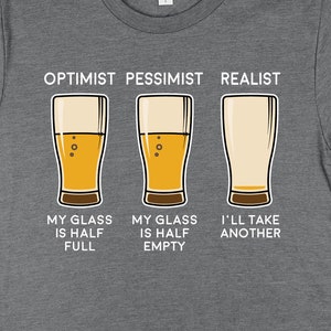 Pode incluir: Camiseta cinza com um gráfico de três copos de cerveja e o texto "Otimista Pessimista Realista". O texto abaixo dos copos diz "Meu copo está meio cheio", "Meu copo está meio vazio" e "Vou tomar outro".