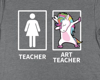 Op de afbeelding: Grijs T-shirt met een witte afbeelding van twee stokfiguren in vakken. Het linker vak zegt "Teacher" en het rechter vak zegt "Art Teacher" met een eenhoorn die de dab doet.