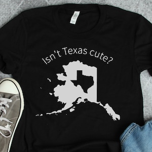 May include: Black t-shirt with a white outline of Alaska with the state of Texas inside. The text "Isn't Texas cute?" is printed on the shirt.