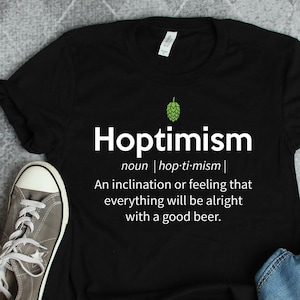 Peut inclure: T-shirt noir avec un graphisme blanc qui dit "Hoptimism" et le définit comme "une inclination ou un sentiment que tout ira bien avec une bonne bière."