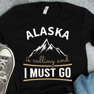 Może przedstawiać: Czarny T-shirt z białym tekstem, który brzmi "Alaska is calling and I must go". Tekst jest otoczony brązową linią. Grafika białego pasma górskiego znajduje się nad tekstem.