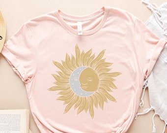 Op de afbeelding: Een roze t-shirt met een gouden zon- en maandesign. De zon heeft een gezicht en de maan heeft een halvemaanvorm.