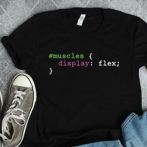May include: Black t-shirt featuring the text "#muscles { display: flex; }" in green and pink. The shirt is styled with grey trainers and denim shorts. The t-shirt is folded, and the trainers are placed on a grey surface.