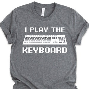 Może przedstawiać: Szara koszulka z napisem "I Play The Keyboard" w kolorze białym, z grafiką pikselowanej klawiatury w kolorze białym.