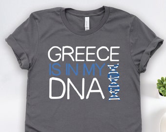 May include: A charcoal grey t-shirt with the text "Greece is in my DNA" in white. A blue and white DNA strand is to the right of the text, with the colors of the Greek flag.
