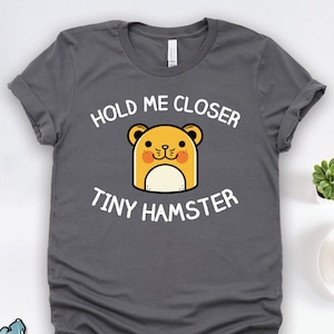 May include: A charcoal grey t-shirt with a white graphic of a cartoon hamster and the text "Hold Me Closer Tiny Hamster".