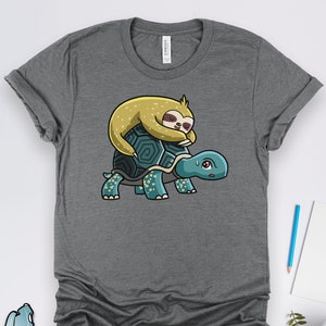 May include: A gray t-shirt with a cartoon illustration of a sloth sleeping on the back of a turtle. The turtle is blue and green with yellow flowers on its shell.