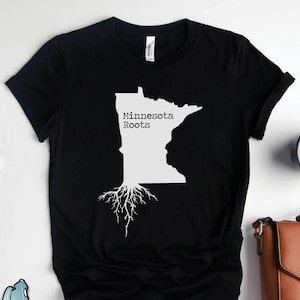 Puede incluir: Camiseta negra con un contorno blanco del estado de Minnesota y el texto "Minnesota Roots" con ra&iacute;ces de &aacute;rbol blancas que se extienden desde la parte inferior del estado.
