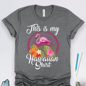 Puede incluir: Camiseta gris con el texto "This is my Hawaiian Shirt" en blanco. Un flamenco rosa con flores tropicales est&aacute; en un c&iacute;rculo detr&aacute;s del texto.