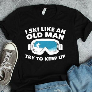 Könnte beinhalten: Schwarzes T-Shirt mit weißem Text, der "I ski like an old man" lautet, und einer Grafik von Skibrillen mit einem Skifahrer im Spiegelbild. Der Text "Try to keep up" befindet sich unter den Brillen.