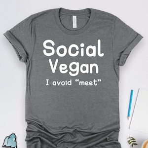 May include: A gray t-shirt with the words "Social Vegan" in large white letters. Below, the text reads "I avoid 'meet'". The shirt is displayed on a white surface with a pen, notepad, and a cup of coffee.