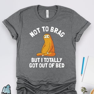 Peut inclure: T-shirt gris avec un dessin animé de paresseux méditant et le texte "Pas pour me vanter mais je suis sorti du lit."