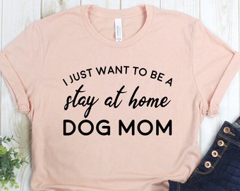 May include: A pink t-shirt with the text "I just want to be a stay at home DOG MOM" in black lettering. The shirt is laid out on a white wooden surface with a pair of white sneakers, blue jeans, and a small green plant.