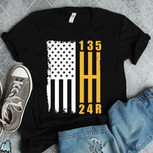 May include: Black t-shirt with a distressed American flag design and a yellow gear shift pattern with the numbers 1, 3, 5, 2, 4, and R.