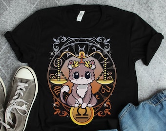 Könnte beinhalten: Schwarzes T-Shirt mit einem Grafikdruck eines Katers mit Krone, der eine Waage hält. Die Waage ist ein Symbol für das Sternzeichen Waage. Die Grafik ist von einem goldenen und braunen, verzierten Design umgeben.