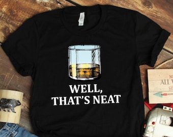 Könnte beinhalten: Schwarzes T-Shirt mit einer Grafik eines Whiskeyglases, gefüllt mit Eis und bernsteinfarbener Flüssigkeit. Der Text "WELL, THAT'S NEAT" ist darunter in Weiß gedruckt. Das Shirt wird auf einer Holzoberfläche präsentiert.