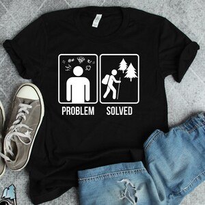 Könnte beinhalten: Schwarzes T-Shirt mit einer weißen Grafik eines Strichmännchens mit einer Gedankenblase über dem Kopf, die mit Symbolen gefüllt ist, die Stress darstellen. Die rechte Seite der Grafik zeigt ein Strichmännchen, das in einem Wald wandert, mit den Worten "Problem gelöst" darunter.
