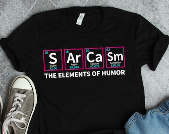 May include: Black t-shirt with a graphic of the periodic table elements, featuring the letters "S", "Ar", "Ca", and "Sm" in pink, with the corresponding element names and atomic weights below. The text "THE ELEMENTS OF HUMOR" is printed below the graphic.