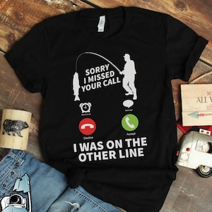 Puede incluir: Camiseta negra con un gráfico blanco de un teléfono con una caña de pescar y un hombre pescando. El texto en la camiseta dice "Lo siento, perdí tu llamada. Estaba en la otra línea."