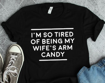 May include: Black t-shirt with white text that reads "I'm so tired of being my wife's arm candy".
