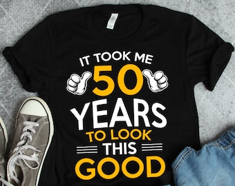 May include: Black t-shirt with yellow text that reads "It took me 50 years to look this good". The text is surrounded by two thumbs up emojis.