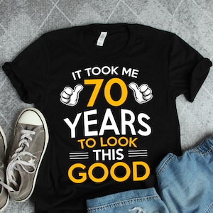 Könnte beinhalten: Schwarzes T-Shirt mit dem Text "Es hat 70 Jahre gedauert, um so gut auszusehen" in Gelb und Weiß. Die Zahl 70 ist groß und von zwei Daumen nach oben umgeben.