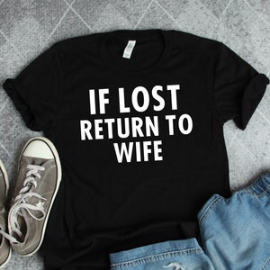 Könnte beinhalten: Schwarzes T-Shirt mit weißem Text, der "IF LOST RETURN TO WIFE" lautet. Das Shirt liegt flach auf einer grauen Oberfläche mit einem Paar grauer Turnschuhe und einer blauen Jeans.