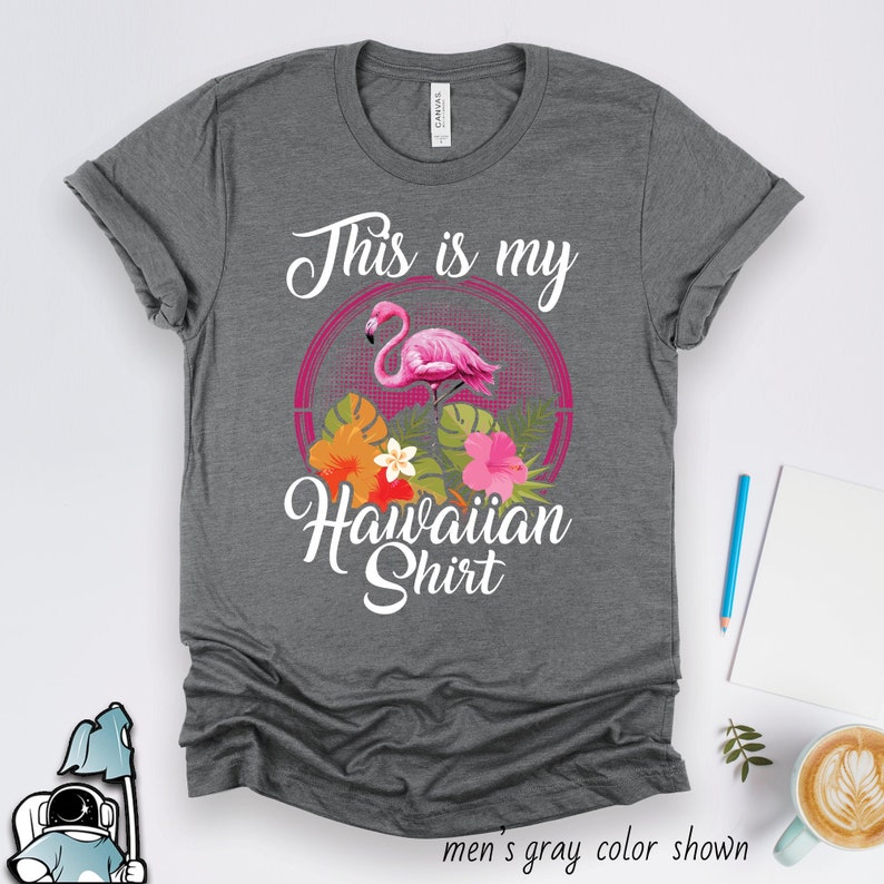 Puede incluir: Camiseta gris con el texto "This is my Hawaiian Shirt" en blanco. Un flamenco rosa con flores tropicales est&aacute; en un c&iacute;rculo detr&aacute;s del texto.