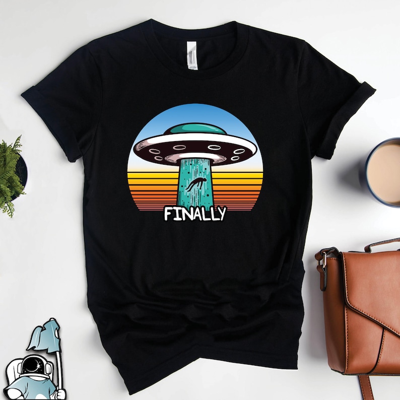 May include: Black t-shirt with a retro-style graphic of a UFO with a person falling out of it. The graphic is set against a sunset background with the text "FINALLY" below it.