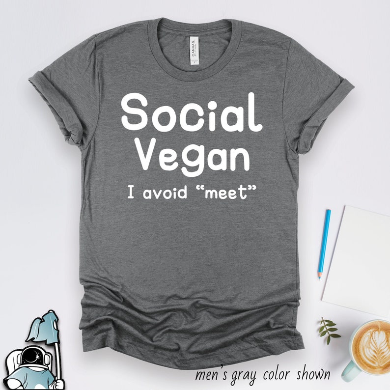 May include: A gray t-shirt with the words "Social Vegan" in large white letters. Below, the text reads "I avoid 'meet'". The shirt is displayed on a white surface with a pen, notepad, and a cup of coffee.